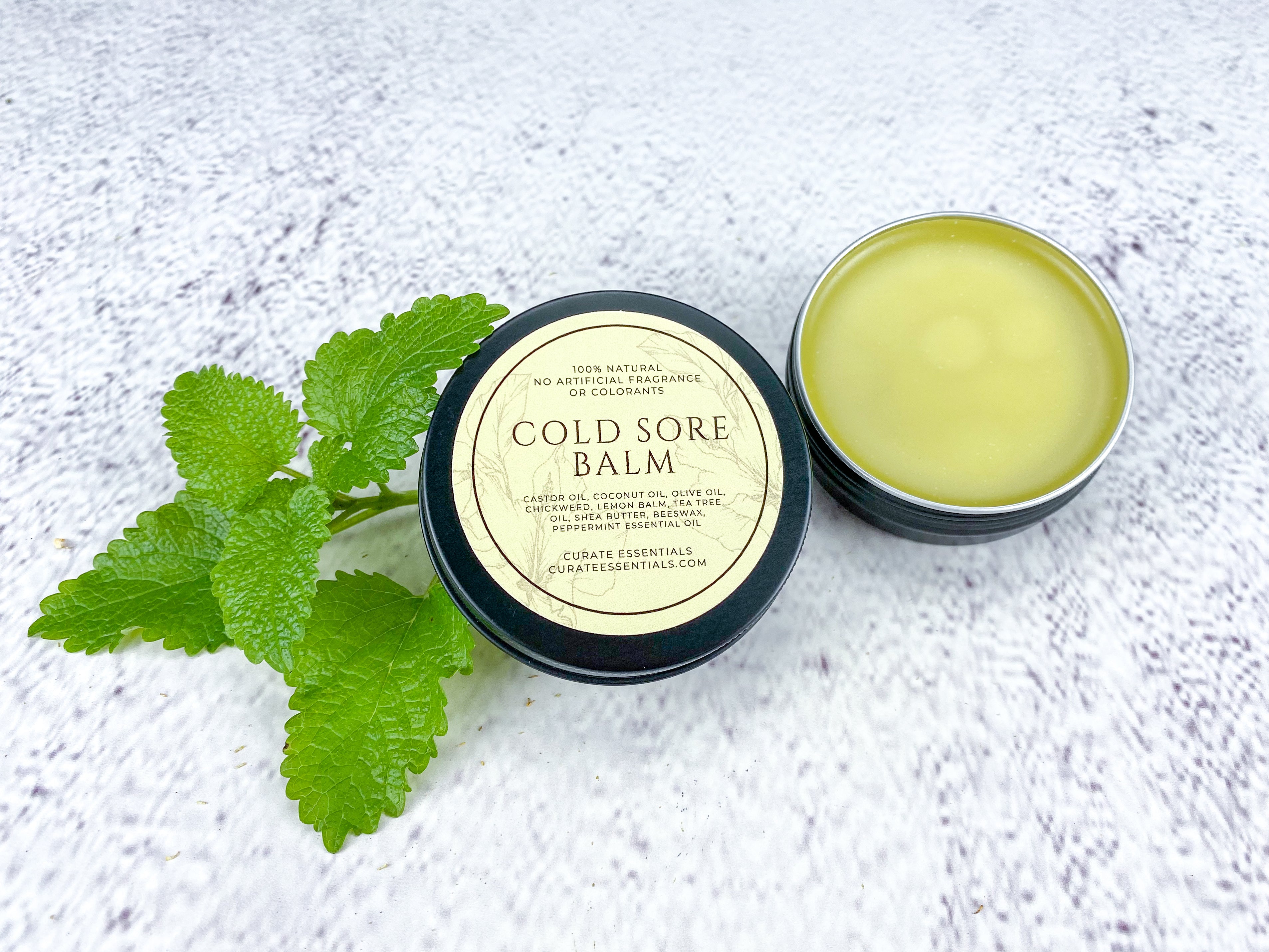 The image showcases a product designed for cold sore relief, presented in a clean, minimalist aesthetic. A small, black, circular tin labeled 'COLD SORE BALM' is the central focus, positioned slightly off-center to the right. The label prominently states '100% NATURAL - NO ARTIFICIAL FRAGRANCE OR COLORANTS.' Beneath this, a list of ingredients is detailed: Castor Oil, Coconut Oil, Olive Oil, Chickweed, Lemon Balm, Tea Tree Oil, Shea Butter, Beeswax, and Peppermint Essential Oil. The brand name, 'CURATE ESSENTIALS,' along with its website 'CURATEESSENTIALS.COM,' appears at the bottom of the label. To the left of the tin, a sprig of vibrant green mint leaves adds a natural, fresh element to the composition. The balm itself, visible inside the open tin, has a pale yellow-green hue. The entire arrangement is set against a textured, white background resembling a marble or tile surface, creating a bright and clinical feel. The lighting is soft and diffused, highlighting the product and its ingredients. The overall impression is one of a natural, effective, and thoughtfully crafted remedy for cold sores, emphasizing purity and quality.