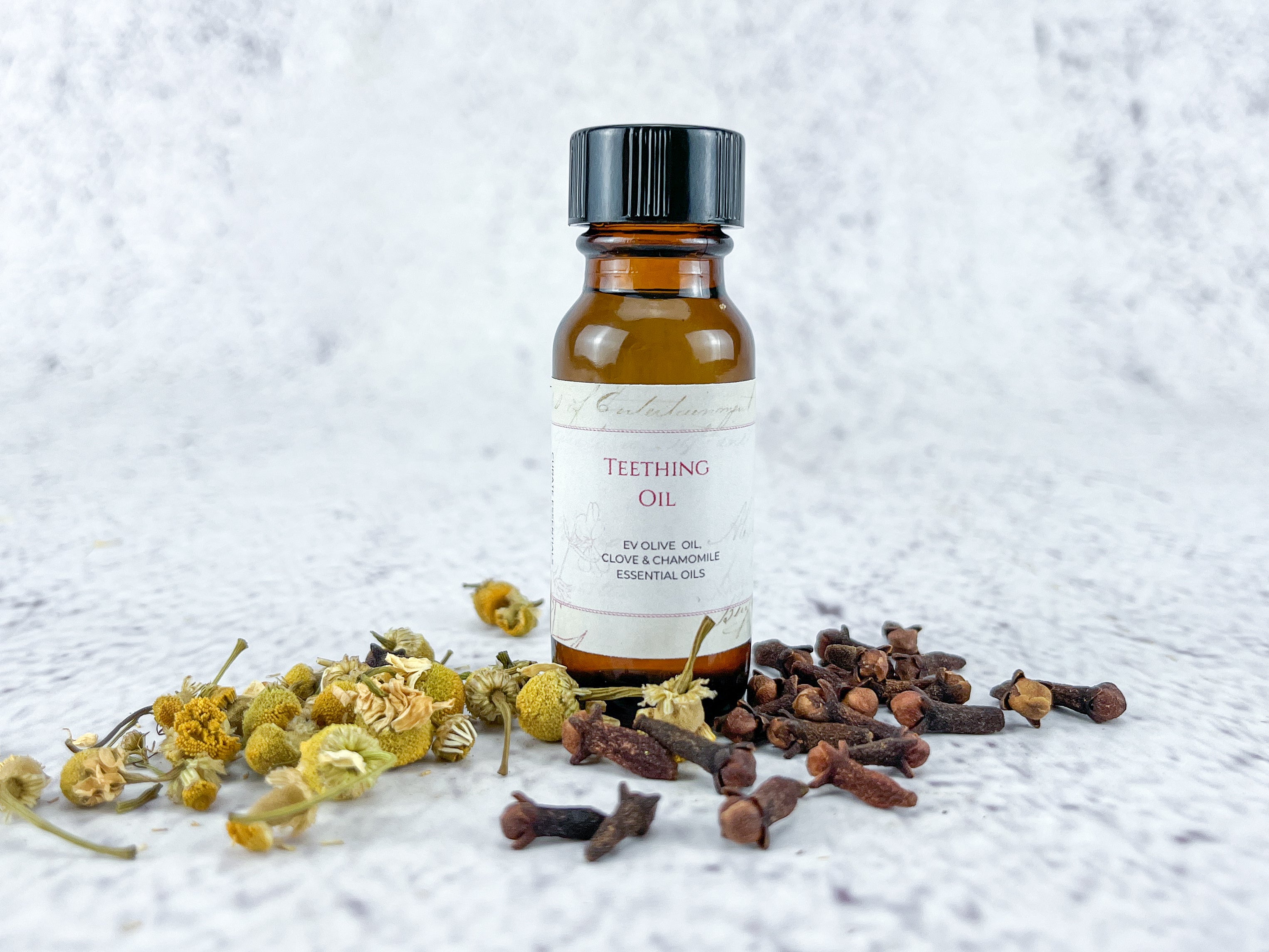 This image showcases a small amber glass bottle of teething oil, positioned centrally against a textured, white and grey marble-like background. The bottle is approximately 1.5 inches tall and has a black screw-on cap. A vintage-style label wraps around the bottle’s midsection, featuring the words 'Teething Oil' in a decorative, slightly faded font. Below this, it specifies the ingredients: 'EV Olive Oil, Clove & Chamomile Essential Oils.' The label has a delicate, ornate border. Surrounding the bottle are scattered natural elements related to the oil’s ingredients. To the left, there’s a cluster of dried chamomile flowers in various shades of yellow and green, some with their stems still attached. On the right, a pile of whole, dark brown cloves are arranged, their distinctive bud-like shape clearly visible. The arrangement feels natural and organic, suggesting a homemade or artisanal product. The lighting is soft and diffused, highlighting the textures of the marble background, the glass bottle, and the dried botanicals. The overall aesthetic is calming and natural, evoking a sense of gentle care and traditional remedies for babies. The composition is well-balanced, drawing the eye to the bottle while the surrounding ingredients reinforce the product’s natural composition.